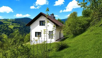 Weißes Einfamilienhaus mit Satteldach auf einem grünen Hügel in ländlicher Umgebung.