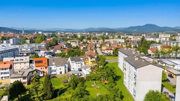 Weitläufige Vogelperspektive einer Stadt mit Wohngebäuden, Grünflächen und Bergen am Horizont.