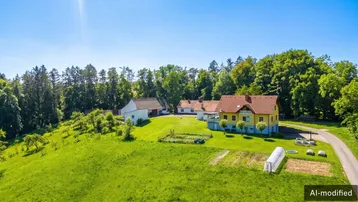 Luftaufnahme eines Anwesens mit mehreren Gebäuden, umgeben von grünen Wiesen und Wäldern unter blauem Himmel.