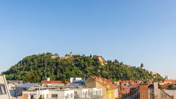 Panoramablick über die Stadt Graz mit dem bewaldeten Schlossberg und dem Uhrturm im Hintergrund.