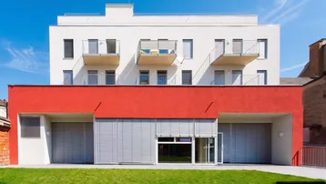 Modernes Wohngebäude mit weißer Fassade, roten Akzenten und kleinen Balkonen über einer Grünfläche.