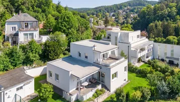 Moderne Villa mit Flachdach und Terrassen, eingebettet in eine grüne Hügellandschaft mit weiteren Häusern.