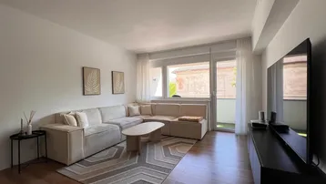 Helles Wohnzimmer mit beigem Ecksofa, Holzboden und Zugang zum Balkon.
