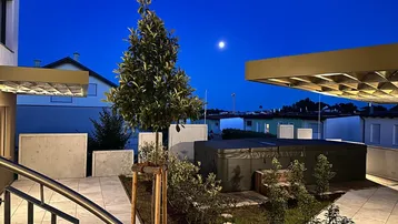Moderne Terrasse mit Whirlpool und Bepflanzung unter dem Nachthimmel, beleuchtet von Außenleuchten.