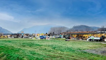 Weitläufige Landschaft mit grüner Wiese im Vordergrund, umgeben von Bergen und einigen Gewerbegebäuden in der Ferne.