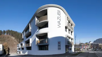 Modernes Gebäude mit geschwungener weißer Fassade, Balkonen und Blick auf bewaldete Hügel und Berge.