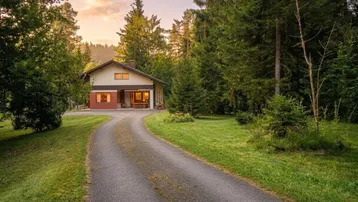 Zufahrt zu einem charmanten Haus, umgeben von hohen Bäumen und grüner Natur bei Sonnenuntergang.