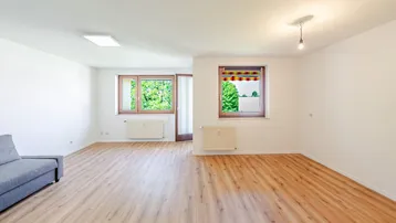 Helles Wohnzimmer mit Laminatboden, weißen Wänden und Fenstern mit Blick ins Grüne.