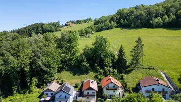 Luftaufnahme einer Wohnsiedlung am Hang, umgeben von Wäldern und grünen Wiesen mit Bergen im Hintergrund.