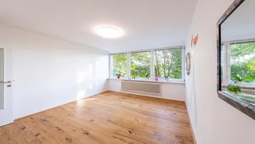 Helles Zimmer mit Holzboden und großen Fenstern, die viel Tageslicht hereinlassen.