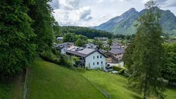 Panoramablick auf eine Wohnsiedlung in einem Tal, umgeben von grünen Hügeln und Bergen.