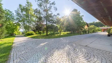 Sonniger gepflasterter Hofbereich mit Bäumen und Blick auf die umliegende Natur.