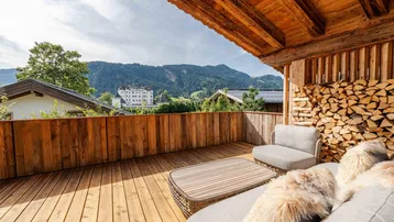 Sonnige Holzterrasse mit bequemen Loungemöbeln und Blick auf die Berge und ein historisches Gebäude.
