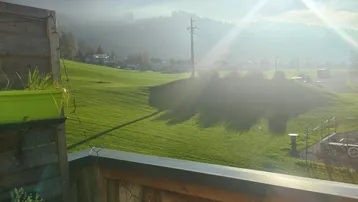 Sonniger Balkon mit Holzelementen und weitem Blick über grüne Wiesen und majestätische Berge.