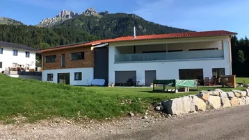 Modernes Haus mit rotem Ziegeldach, weißer Fassade und Holzverkleidung vor einer beeindruckenden Bergkulisse.