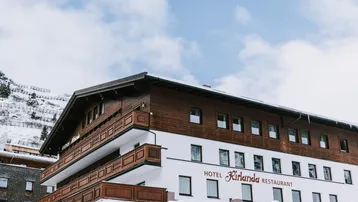 Ansicht des Hotels Kirlana mit traditioneller Holzfassade und schneebedeckten Bergen im Hintergrund.