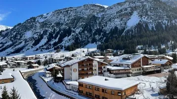 Panoramablick auf ein verschneites Bergdorf mit traditionellen Gebäuden und einem Flusslauf unter strahlend blauem Himmel.