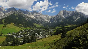 Panoramablick auf ein malerisches Dorf in einem Tal, umgeben von majestätischen Bergen und grünen Wiesen.
