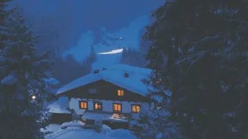 Traditionelles Chalet im Schnee bei Nacht, umgeben von verschneiten Bäumen und Bergen.