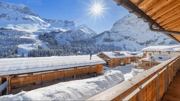 Panoramablick vom Balkon auf schneebedeckte Berge und traditionelle Chalets unter strahlendem Sonnenschein.