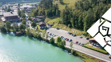 Luftaufnahme eines Dorfes am Seeufer mit Parkplätzen und bewaldeten Bergen im Hintergrund.