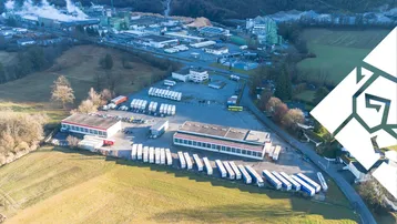 Luftaufnahme eines großen Gewerbegebiets mit mehreren Hallen, LKW-Parkplätzen und einer Fabrik im Hintergrund.