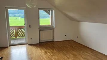 Helles Zimmer im Dachgeschoss mit Holzboden und Zugang zum Balkon, bietet einen weiten Blick ins Grüne.