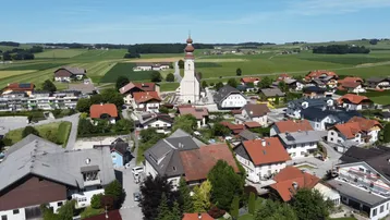 Idyllische Luftaufnahme eines Dorfes mit zentraler Kirche, umgeben von weitläufigen grünen Feldern unter blauem Himmel.