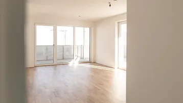 Helles, leeres Zimmer mit Holzboden und Zugang zu einem Balkon, durchflutet von Tageslicht.