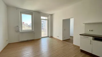 Helles Wohnzimmer mit Laminatboden, Zugang zum Balkon und offener Küchenzeile.
