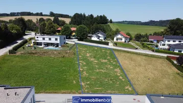 Luftaufnahme eines Baugrundstücks mit blauer Umrandung, umgeben von modernen Häusern und grüner Landschaft.