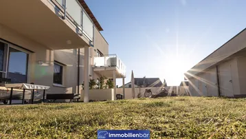 Modernes Gebäude mit Balkonen und einer sonnigen Rasenfläche im Vordergrund, bei klarem Himmel.