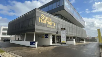 Modernes Bürogebäude mit Glasfassade und Parkplätzen unter einem bewölkten Himmel, das eine professionelle Geschäftsadresse bietet.
