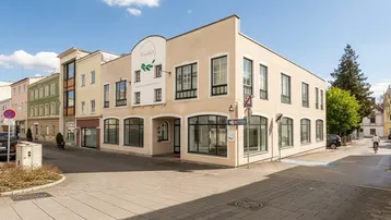 Das Eckgebäude präsentiert sich mit großen Schaufenstern und einer hellen Fassade an einer belebten Straße.