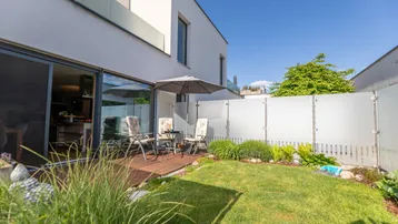 Moderne Terrasse mit Holzdeck, Gartenmöbeln und kleinem Rasenbereich vor einem weißen Gebäude.