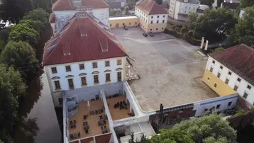 Luftaufnahme eines historischen Gebäudekomplexes mit großem Innenhof und einer Terrasse am Wasser.