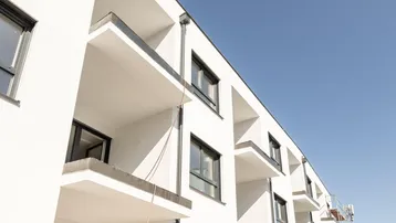 Moderne weiße Fassade eines Neubaus mit Balkonen unter klarem blauen Himmel.
