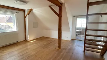 Heller Dachgeschoss-Wohnbereich mit sichtbaren Holzbalken, Holztreppe und natürlichem Lichteinfall durch Fenster.