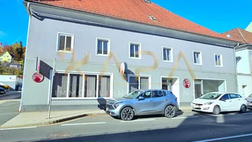 Graue Fassade eines Mehrfamilienhauses mit rotem Ziegeldach und parkenden Autos davor.