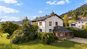 Weißes Einfamilienhaus mit großem Garten und grüner Umgebung unter blauem Himmel.