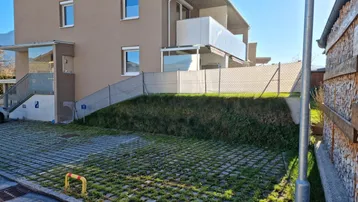 Modernes Mehrfamilienhaus mit Balkonen, begrüntem Parkplatzbereich und Zugang über eine Rampe.