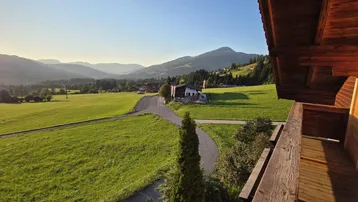 Panoramablick von einem Holzbalkon über grüne Wiesen und sanfte Berge bei Sonnenuntergang.