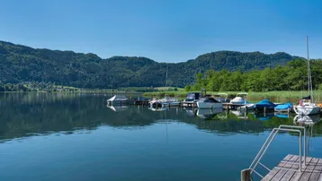 Idyllischer See mit Bootsstegen und bewaldeten Bergen im Hintergrund unter blauem Himmel.