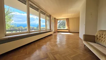 Geräumiges Wohnzimmer mit Fischgrätparkett, großen Fenstern und weitem Blick auf die Berge und Natur.