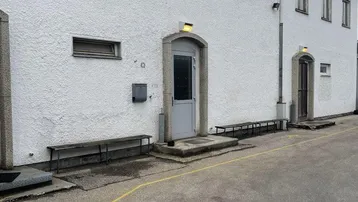 Weißes Gebäude mit mehreren Eingängen und Fenstern, davor ein asphaltierter Bereich mit gelben Markierungen.