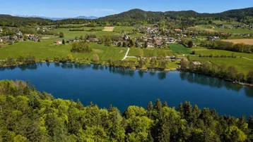 Malerische Luftaufnahme eines Sees umgeben von grünen Hügeln und einem Dorf.