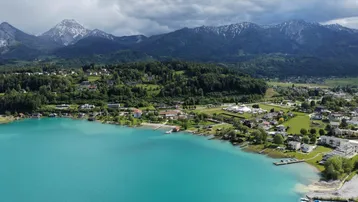 Luftaufnahme eines türkisfarbenen Sees mit angrenzender Ortschaft und schneebedeckten Bergen im Hintergrund.