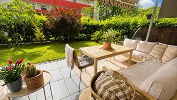 Möblierte Terrasse mit Markise, umgeben von üppigem Grün und Blick auf das Gebäude.