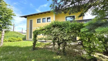 Gelbes Haus mit grüner Rasenfläche und üppiger Vegetation unter blauem Himmel.
