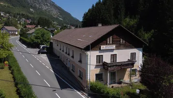 Luftaufnahme eines traditionellen Gasthauses an einer Straße, umgeben von grünen Bergen und Wäldern.